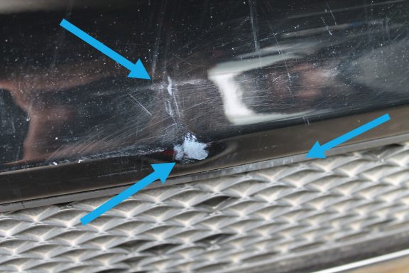 Lackabplatzer mit verformtem Material Kratzer und Dellen auf einer schwarzen Fahrzeugoberfläche mit sichtbaren Pfeilen.