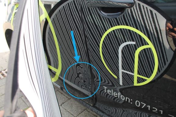 Seitenschaden Verformung mit Dellensegel Schwarze Autotür mit gelben und blauen Designelementen und Kontaktnummer.
