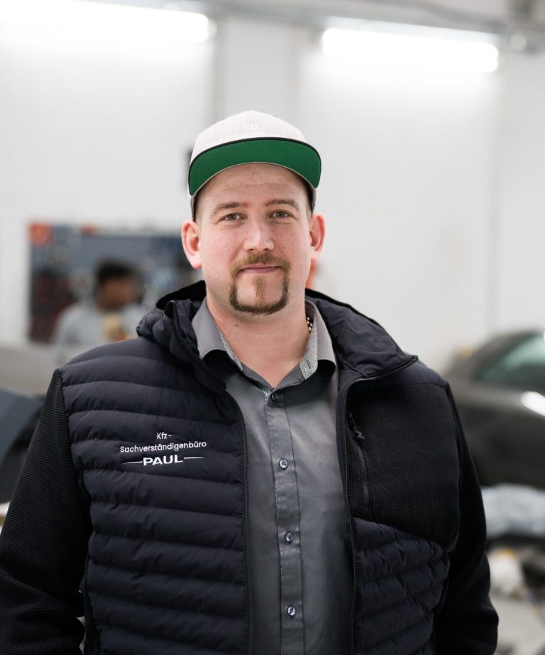 Jens Paul - Inhaber des Kfz-Sachverständigenbüro Paul Mann mit Hut und Weste, lächelt in einem modernen Innenraum.