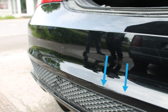 Schadenübersicht Nahaufnahme von einem schwarzen Autokofferraum mit zwei Pfeilen, die auf eine Struktur zeigen.