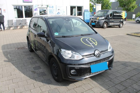 VW UP Seitenschaden rechts Schwarzer Kleinwagen mit gelbem Logo, parkt auf einem Asphaltplatz.